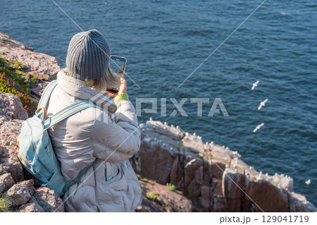 Woman in warm clothing sitting on a rocky Arctic cliff at the Barents Sea in Teriberka park, photographing a seabird colony with her smartphone 129041719