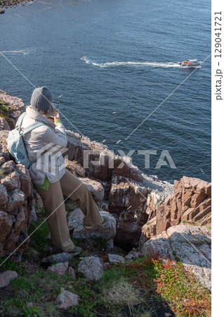 Woman with backpack and winter clothing standing on a rocky cliff by the Barents Sea in Teriberka, photographing a boat and seabird colony below 129041721