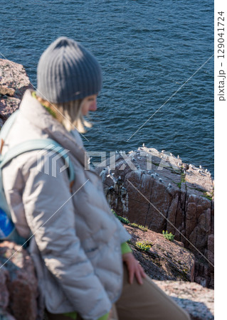 Woman standing on a rocky Arctic cliff at the Barents Sea, observing a seabird colony nesting on red coastal rocks 129041724