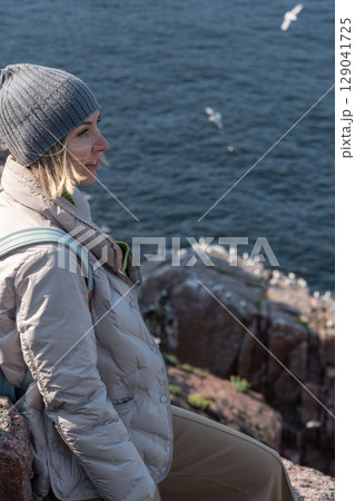 Smiling woman in a gray beanie and light down jacket standing on a rocky Arctic cliff above the Barents Sea, observing a seabird colony below 129041725