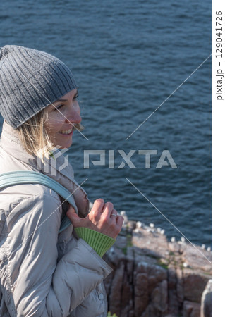 Smiling woman in a gray beanie and light down jacket standing on a rocky Arctic cliff above the Barents Sea, observing a seabird colony below 129041726