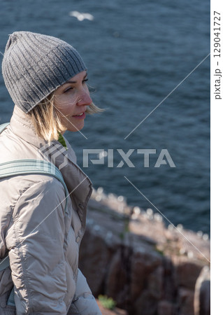 Smiling woman in a gray beanie and light down jacket standing on a rocky Arctic cliff above the Barents Sea, observing a seabird colony below Smiling woman in a gray beanie and light down jacket standing on a rocky Arctic cliff above the Barents Sea, observing a seabird colony below 129041727