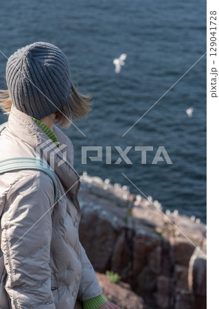 A woman in a gray beanie and light jacket stands on a rocky cliff by the Barents Sea, gazing at the flying seabirds near a nesting colony 129041728