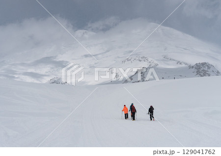 Group of climbers with trekking poles and backpacks ascending snow-covered Mount Elbrus under dramatic cloudy skies Group of climbers with trekking poles and backpacks ascending snow-covered Mount Elbrus under dramatic cloudy skies 129041762