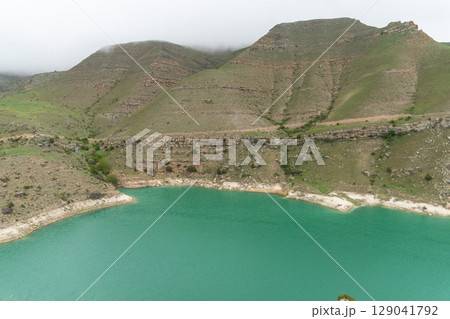 A vivid turquoise mountain lake winds through the lush, mist-covered hills of the North Caucasus 129041792
