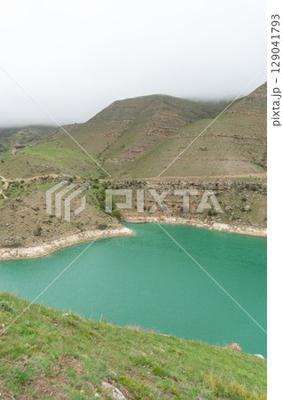 A vivid turquoise mountain lake winds through the lush, mist-covered hills of the North Caucasus 129041793
