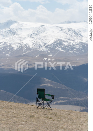Solitary green camping chair stands on a mountain slope facing the majestic snow-covered peaks of the Caucasus Mountains 129041830