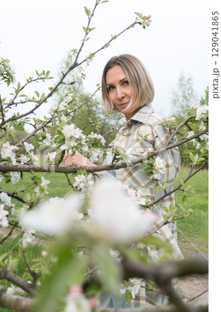 Woman in a plaid coat standing among blooming branches in a spring orchard, softly gazing through apple blossoms. Femininity, self-connection, beauty in nature, and spring renewal Woman in a plaid coat standing among blooming branches in a spring orchard, softly gazing through apple blossoms. Femininity, self-connection, beauty in nature, and spring renewal 129041865