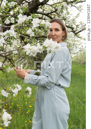 Middle-aged woman in soft blue loungewear standing among blooming apple trees. Wellness retreats, organic skincare, spring fashion, and mental health campaigns can all benefit from this serene 129041887