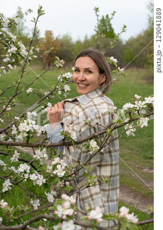 Woman in a plaid coat standing among blooming branches in a spring orchard, softly gazing through apple blossoms. Femininity, self-connection, beauty in nature, and spring renewal Woman in a plaid coat standing among blooming branches in a spring orchard, softly gazing through apple blossoms. Femininity, self-connection, beauty in nature, and spring renewal 129041889