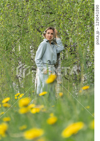 Middle-aged woman in a soft blue outfit standing in a birch grove with blooming dandelions in the foreground. Natural lifestyle, beauty in simplicity, and branding for eco cosmetics, wellness retreats 129041890