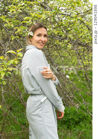 Confident middle-aged woman in light blue clothing smiling in a spring orchard. Personal care, skincare, and wellness industries may use this image to convey natural beauty and authenticity Confident middle-aged woman in light blue clothing smiling in a spring orchard. Personal care, skincare, and wellness industries may use this image to convey natural beauty and authenticity 129041898