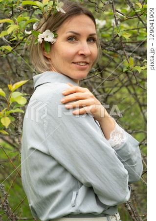 Confident middle-aged woman with a flower tucked in her hair standing among tree branches. Wellness brands, skincare advertising, and campaigns focused on natural aging 129041909