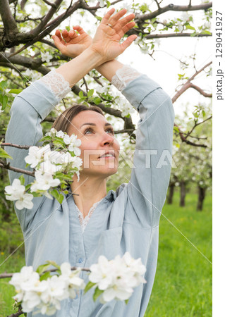 A middle-aged woman in a pale blue outfit stands under blossoming apple trees with her arms raised gracefully. Inner peace, holistic wellness, feminine energy A middle-aged woman in a pale blue outfit stands under blossoming apple trees with her arms raised gracefully. Inner peace, holistic wellness, feminine energy 129041927