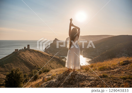 woman stands on a hill overlooking the ocean, her arms raised in the air. Concept of freedom and joy, as if the woman is celebrating a moment of happiness or accomplishment. 129043833