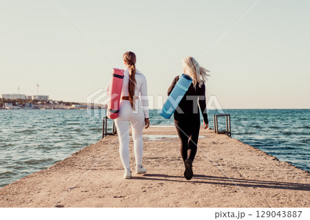 Yoga Women Beach Pier - Two women walk on a pier toward the ocean carrying yoga mats, enjoying a sunrise or sunset. 129043887
