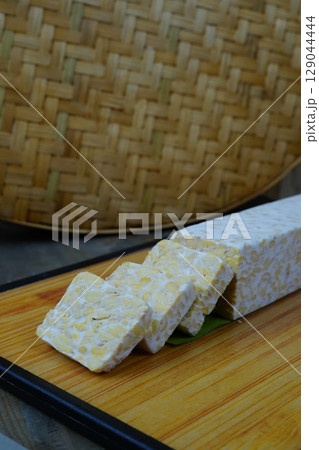 A block of tempeh, partially sliced, sits on a wooden cutting board with a woven bamboo mat in the background. A block of tempeh, partially sliced, sits on a wooden cutting board with a woven bamboo mat in the background. 129044444