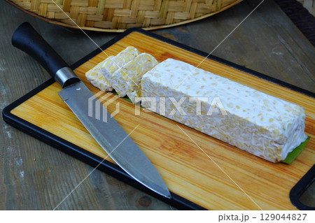 A block of tempeh, partially sliced, on a wooden cutting board with a knife, set against a woven bamboo background. 129044827