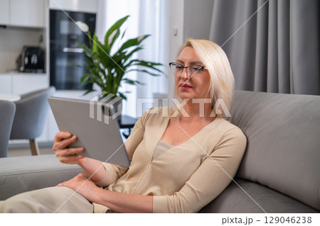 Smiling middle aged lady enjoying a film on her tablet, seated on a cozy sofa at home. Entertainment, leisure and modern technology concept. 129046238