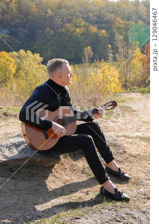 Blonde man playing string guitar outdoors in autumn forest. Concept of sound therapy, mental health and wellness rituals. Calmness tranquility audio-sensory practices. Aura farming energy 129046467