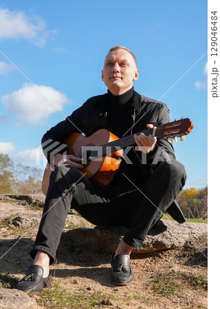 Beautiful blonde man playing acoustic guitar and singing relaxing enjoying outdoors on sunny autumn day. Artist learning and practicing playing musical instrument. Fall leaves autumnal 129046484
