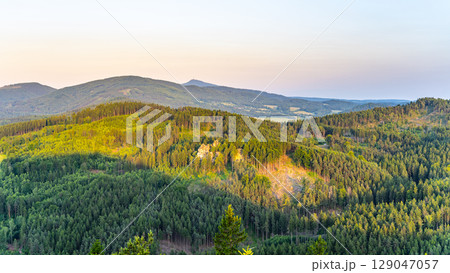 Jested Ridge is bathed in golden light as the sun sets behind the Lusatian Mountains. The sandstone cliffs rise above lush green forests, creating a breathtaking evening view. 129047057