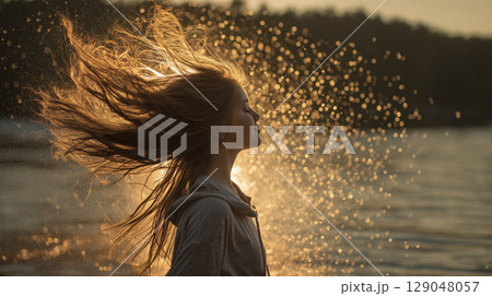 Woman with flowing hair stands near body of water during golden hour, with sunlight creating sparkling effect behind her and serene, peaceful atmosphere Woman with flowing hair stands near body of water during golden hour, with sunlight creating sparkling effect behind her and serene, peaceful atmosphere 129048057