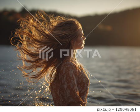 Woman with long hair flipping in wind near body of water during sunset, creating dynamic and serene scene with warm lighting and natural surroundings 129048061