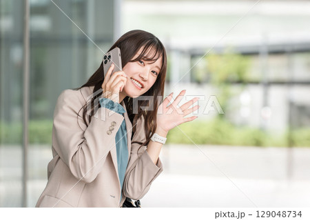 都会のオフィス街でスマホで通話しながら手を振る若いアジア人ビジネスウーマン（待ち合わせ） 129048734