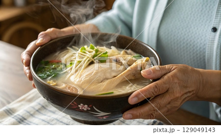 Samgyetang held in the hands of a Korean grandmother wearing traditional hanbok in a warmly lit home kitchen environment 129049143