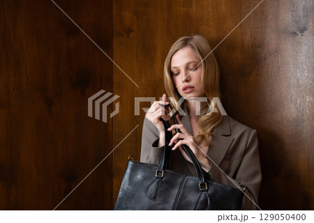 A stylish young woman, long blonde hair, poses in a grey suit, holding a black leather bag against a warm wooden background 129050400