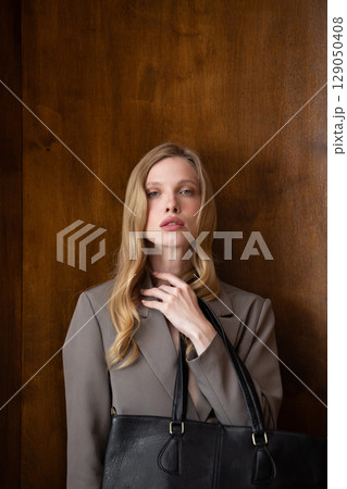 A stylish young woman, long blonde hair, poses in a grey suit, holding a black leather bag against a warm wooden background 129050408
