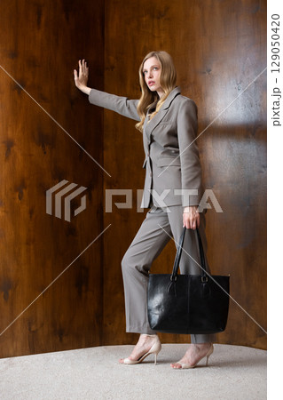 A stylish young woman, long blonde hair, poses in a grey suit, holding a black leather bag against a warm wooden background 129050420