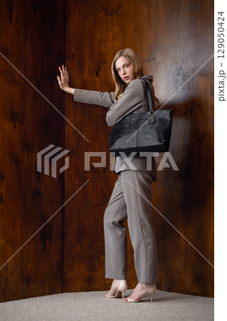 A stylish young woman, long blonde hair, poses in a grey suit, holding a black leather bag against a warm wooden background 129050424