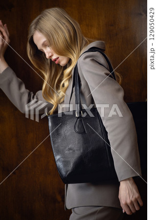 A stylish young woman, long blonde hair, poses in a grey suit, holding a black leather bag against a warm wooden background 129050429