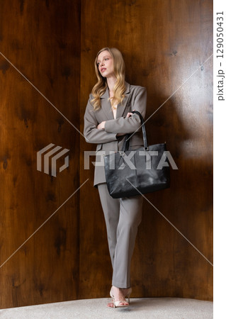 A stylish young woman, long blonde hair, poses in a grey suit, holding a black leather bag against a warm wooden background 129050431