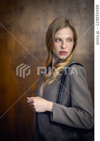 A stylish young woman, long blonde hair, poses in a grey suit, holding a black leather bag against a warm wooden background 129050489