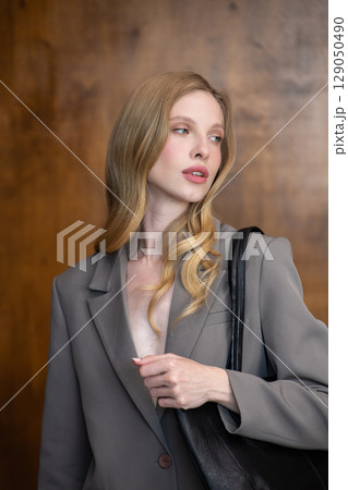 A stylish young woman, long blonde hair, poses in a grey suit, holding a black leather bag against a warm wooden background 129050490