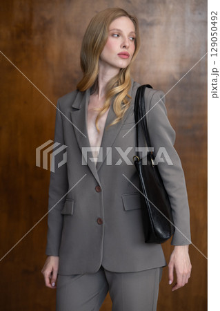 A stylish young woman, long blonde hair, poses in a grey suit, holding a black leather bag against a warm wooden background 129050492