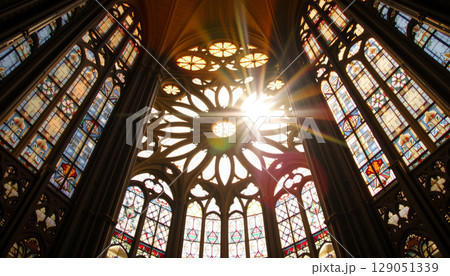 Stained glass window with sun rays shining through intricate gothic cathedral architecture creating warm glowing effect Stained glass window with sun rays shining through intricate gothic cathedral architecture creating warm glowing effect 129051339