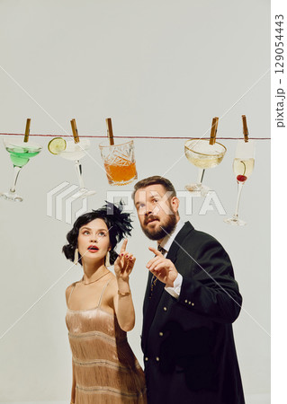 Man and woman in retro party outfits pointing at cocktails clipped to string with surprised faces 129054443