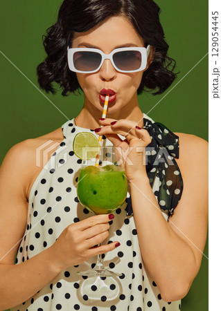 Woman in polka-dot dress sipping green drink with lime through striped straw on green background Woman in polka-dot dress sipping green drink with lime through striped straw on green background 129054445