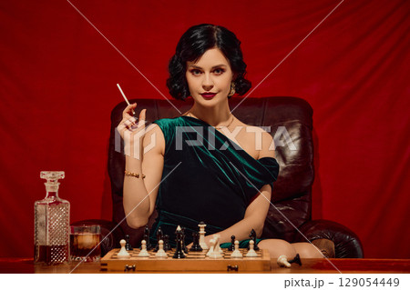 Woman in velvet dress holding cigarette and glass sitting at chessboard with intense expression 129054449