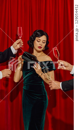 Woman in velvet dress pouring champagne from bottle into glass while surrounded by raised hands Woman in velvet dress pouring champagne from bottle into glass while surrounded by raised hands 129054452