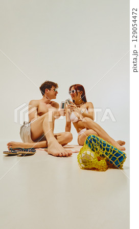 Happy young couple sitting on towel with cans and beach accessories smiling at each other Happy young couple sitting on towel with cans and beach accessories smiling at each other 129054472