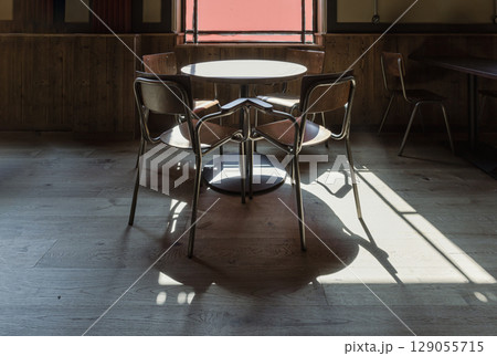 Sunlight streams through large window onto an the wooden dining tables, chairs and the floor with dark shadows in the background. Sunlight streams through large window onto an the wooden dining tables, chairs and the floor with dark shadows in the background. 129055715