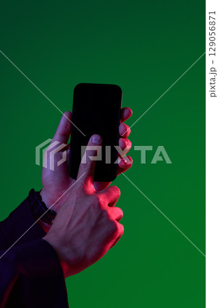 Hands holding smartphone against vibrant green and red lighting background 129056871