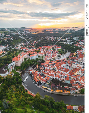 The sun rises over Cesky Krumlov, casting a warm glow on the charming town's rooftops and the winding Vltava River. UNESCO heritage sites and lush landscapes create a picturesque view. 129058066