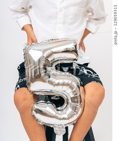 Close-up of a young boy holding a shiny silver number 5 balloon, wearing casual summer outfit, celebrating birthday indoors Close-up of a young boy holding a shiny silver number 5 balloon, wearing casual summer outfit, celebrating birthday indoors 129059313