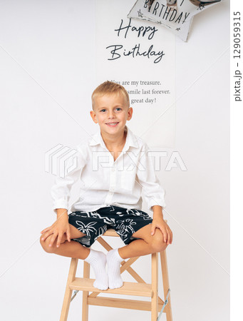 Happy young boy in casual clothes sitting on a wooden stool in front of a birthday greeting sign and balloon Happy young boy in casual clothes sitting on a wooden stool in front of a birthday greeting sign and balloon 129059315